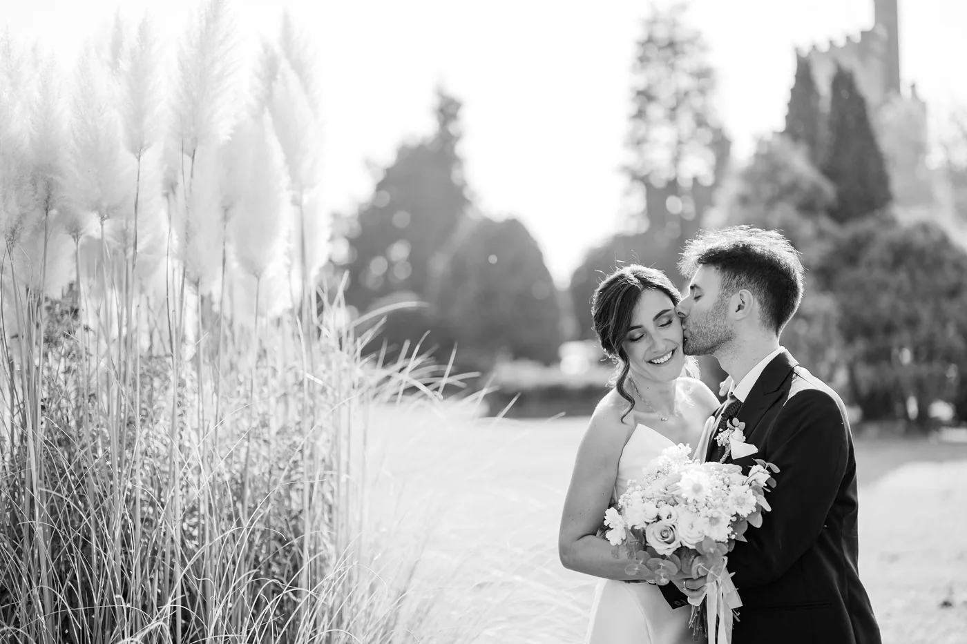 Reviews 1 The groom kisses the smiling bride on the cheek beside pampas grass, in black and white